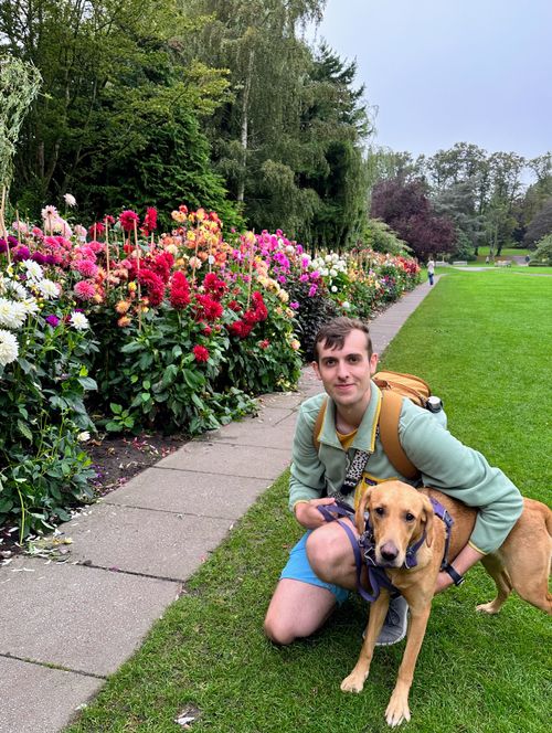 Joe crouched, hugging Betsty, both stood alongside rows of dahlias. The colours of the dahlias range from groups of bright whites, deep reds, neon pinks, and some a mix of orange and red resembling a sunset.