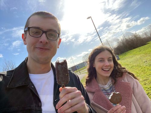 Myself and Naomi holding up ice creams and smiling with a bright blue sky in the background. I am wearing a white top with a black denim jacket, my glasses. Naomi is wearing a suede looking material pink jacket. In the background are the leafless branches from a bush in the midst of bright green grass. The sky is a vivid blue with clouds strewn about, making the lighting indirect, albeit bright.