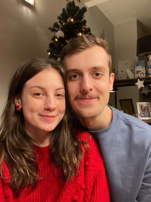 Myself and Naomi taking a selfie, with me smiling and wearing a light blue jumper and Naomi wearing a red knitted jumper and festive earrings - both smiling, with our Christmas tree, covered in small white lights, visible in the background.