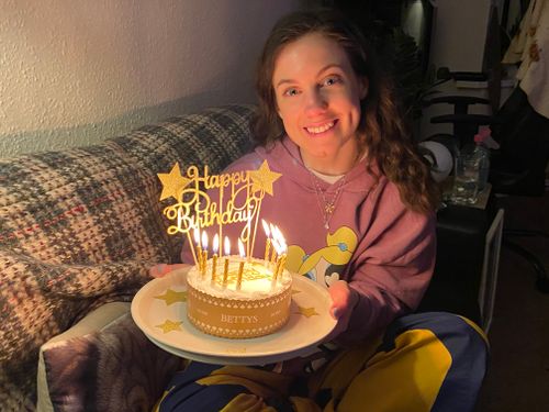 My partner Naomi, wearing her new oversized navy and mustard coloured trousers and purple hoodie featuring all of the Powerpuff Girls as well a big smile. Naomi is holding a small tray with a white iced Betty’s ‘Happy Birthday’ cake. The cake features a small banner that says: Happy Birthday. In front of the banner are 10 small candles. Naomi is sitting on the sofa, with a cosy blanket on the back, in the background are the beige coloured walls of our front room and our ZZ plant sitting on a small coffee table behind the sofa.