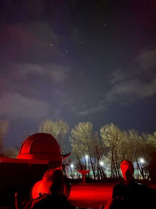 The dark blue night sky takes up the majority of the photo, with bright white stars visible, covered by whispy thin clouds. In the foreground, red lights illuminate a demonstrator and some walls, in the background is a 2 storey observatory which sits in front of a row of tall thin trees.