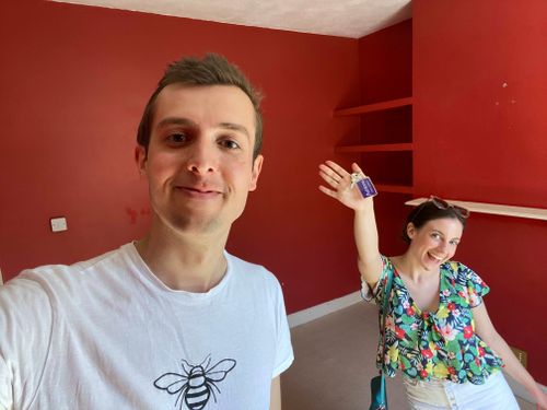 Myself and Naomi stood in a deep red room, with Naomi smiling and holding the keys to our new house above her head. In the background are some plain red walls and empty shelves.