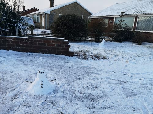 A small snowperson (roughly 20cm tall) sits at the front of the image with two stick arms, coal eyes, a coal nose and three coal buttons. The ground is covered in a thin blanket of snow. In the background is the snowperson we built previously. The rest of the background is made up by a small brick wall surrounding some grass, a series of bushes and our bungalow. 