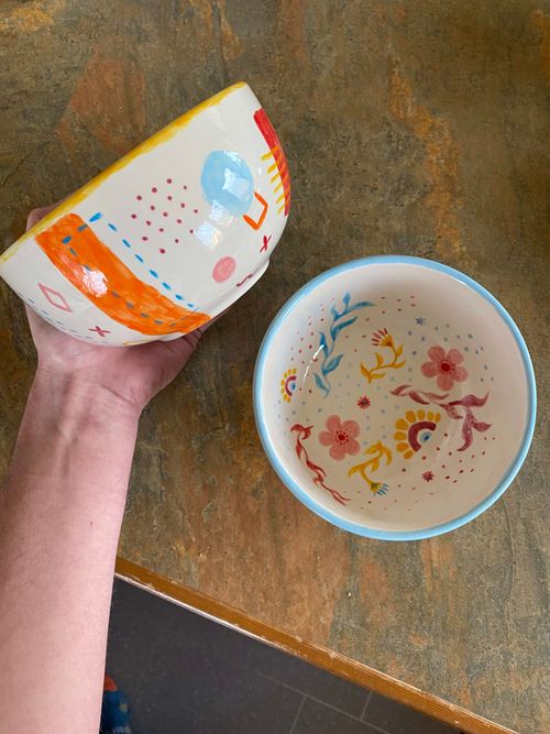 Two glossy, painted bowls. With my left hand I am holding a bowl that I painted featuring orange, blue and pink shapes - some filled in, some with just their outlines. The rim of the bowl is painted a bright yellow colour. In the photo, the side of the bowl can be seen. To the right of the photo is the bowl that Naomi painted. You can only see the inside which has been patterned with yellow and pink flowers with blue, yellow and red vines surrounding them. The rim of the bowl is painted a light sky blue colour. The background of the photo is our drab green brown worktops.